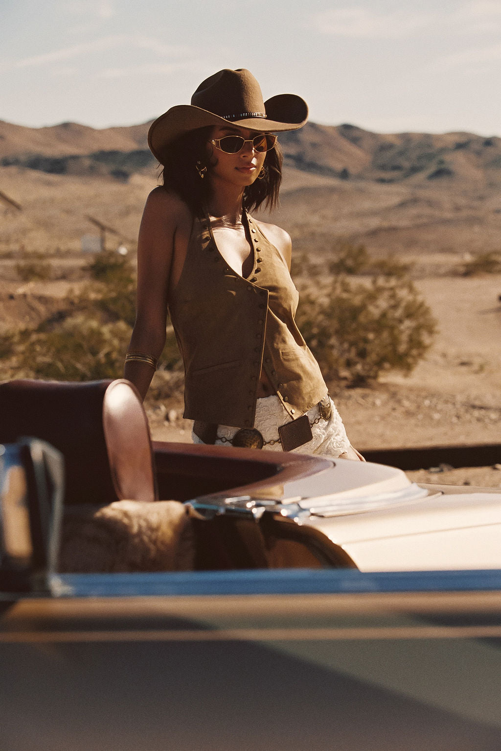 Thatcher Camel Suede Studded Top and Posie Ivory Floral Micro Shorts styled with the Dune Coin Statement Belt and cowboy hat leaning against a vintage convertible in the desert.