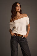 Front view of a woman modeling a cream asymmetrical sweater with dark jeans and a belt