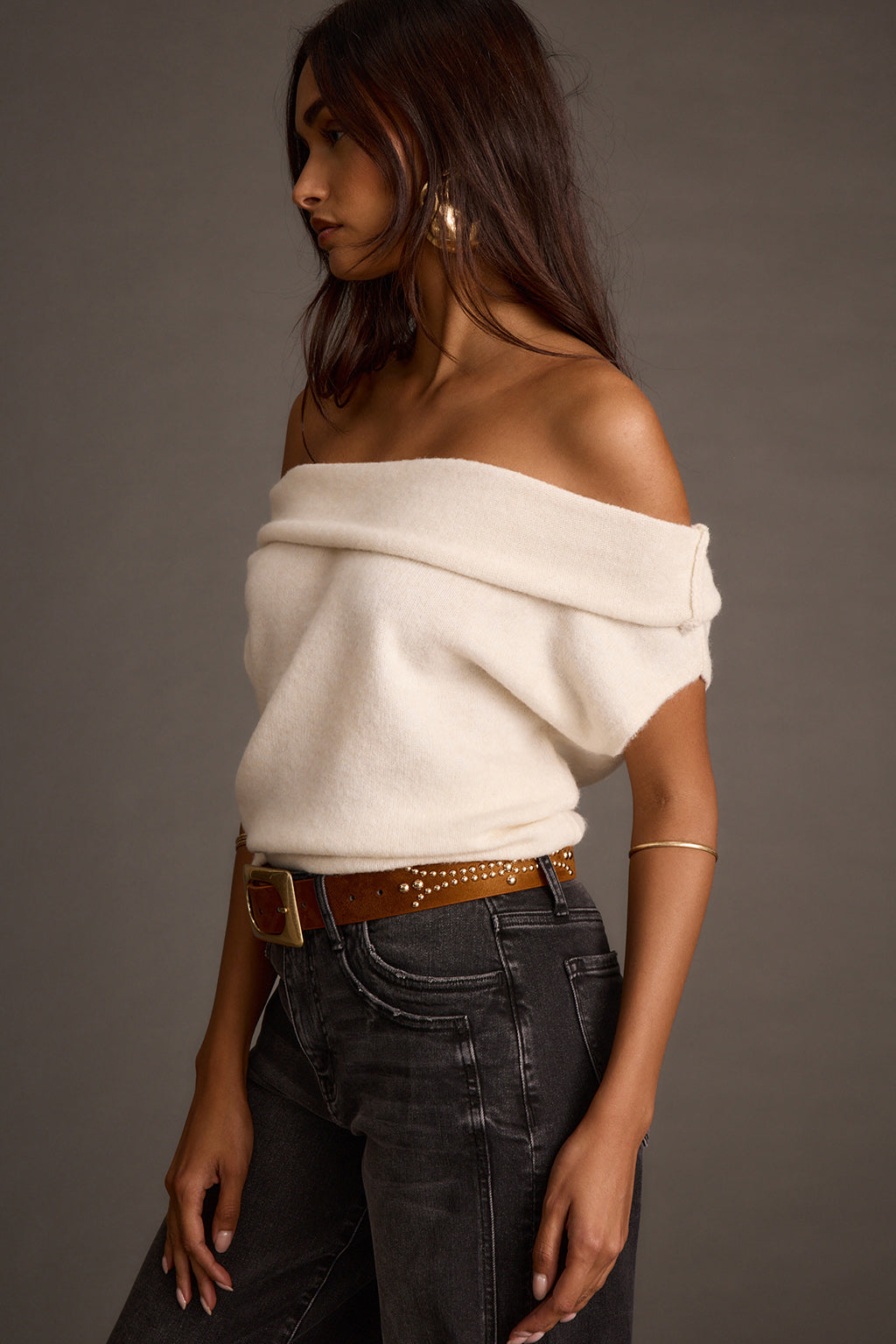 Side profile of a cream asymmetrical sweater with a studded belt and dark jeans