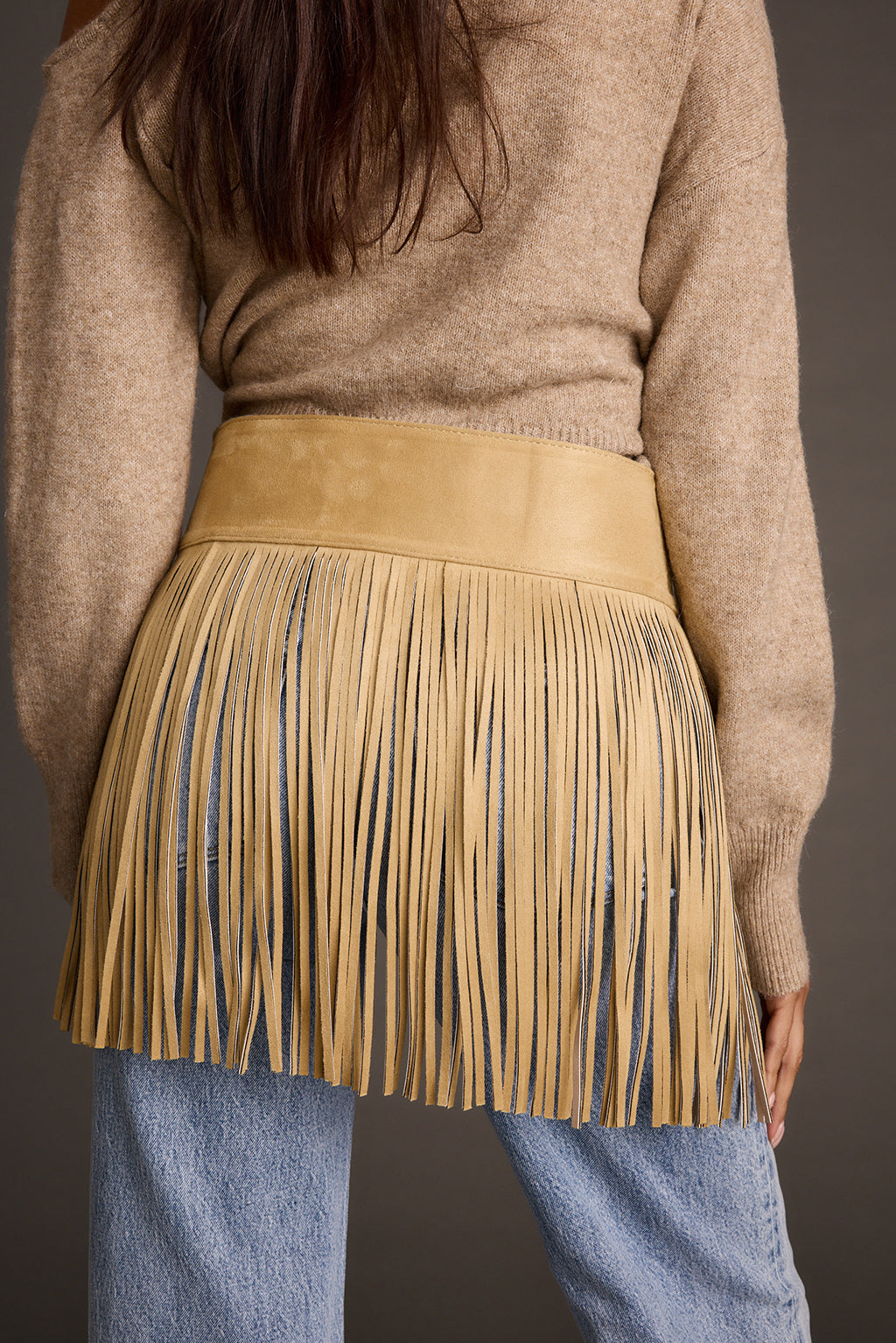 Close-up back view of a camel faux suede fringe belt worn over a beige sweater and jeans
