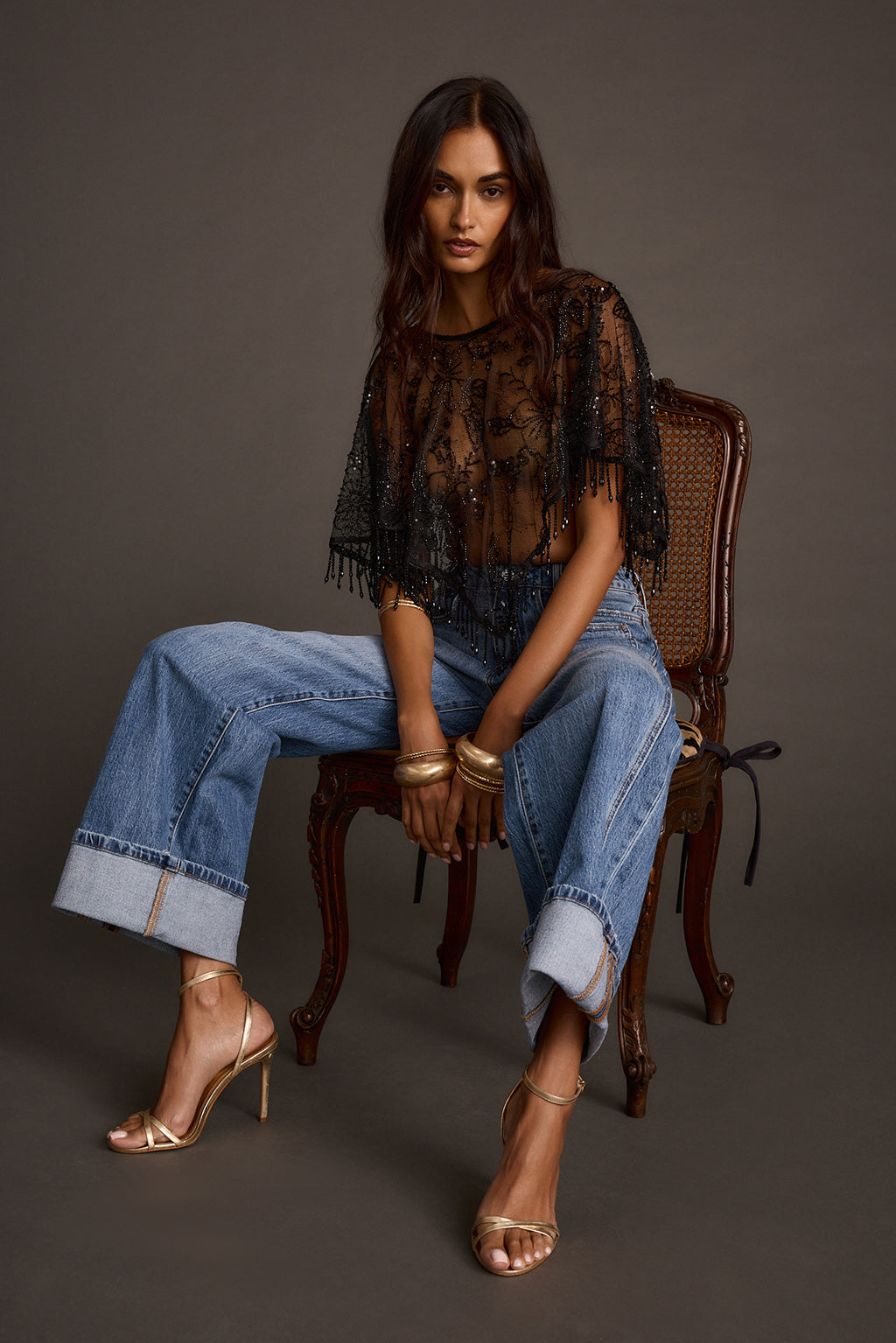 Woman seated wearing a sheer black fringe shawl top with wide-leg jeans and gold heels