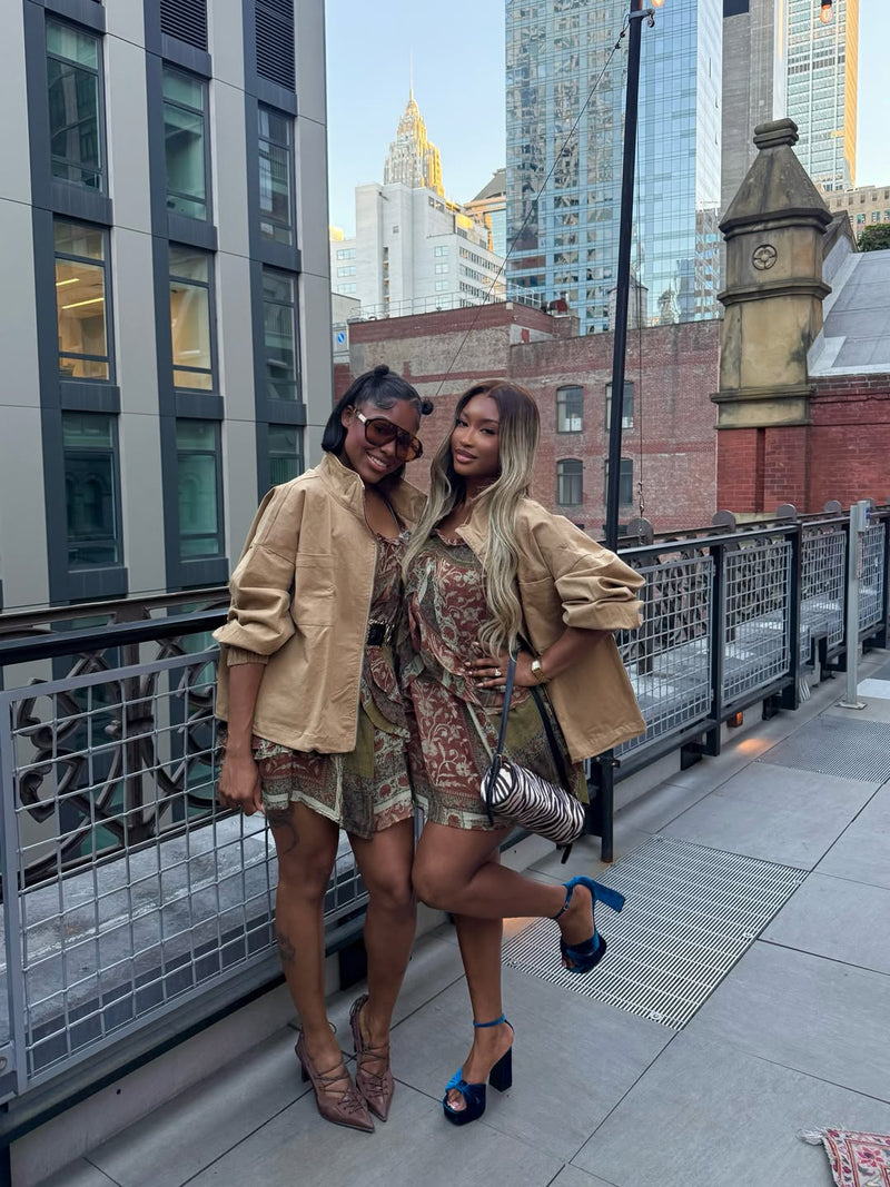 Two women wearing beige trench coats over patterned mini dresses, posing on an elevated city walkway with the skyline in the background.