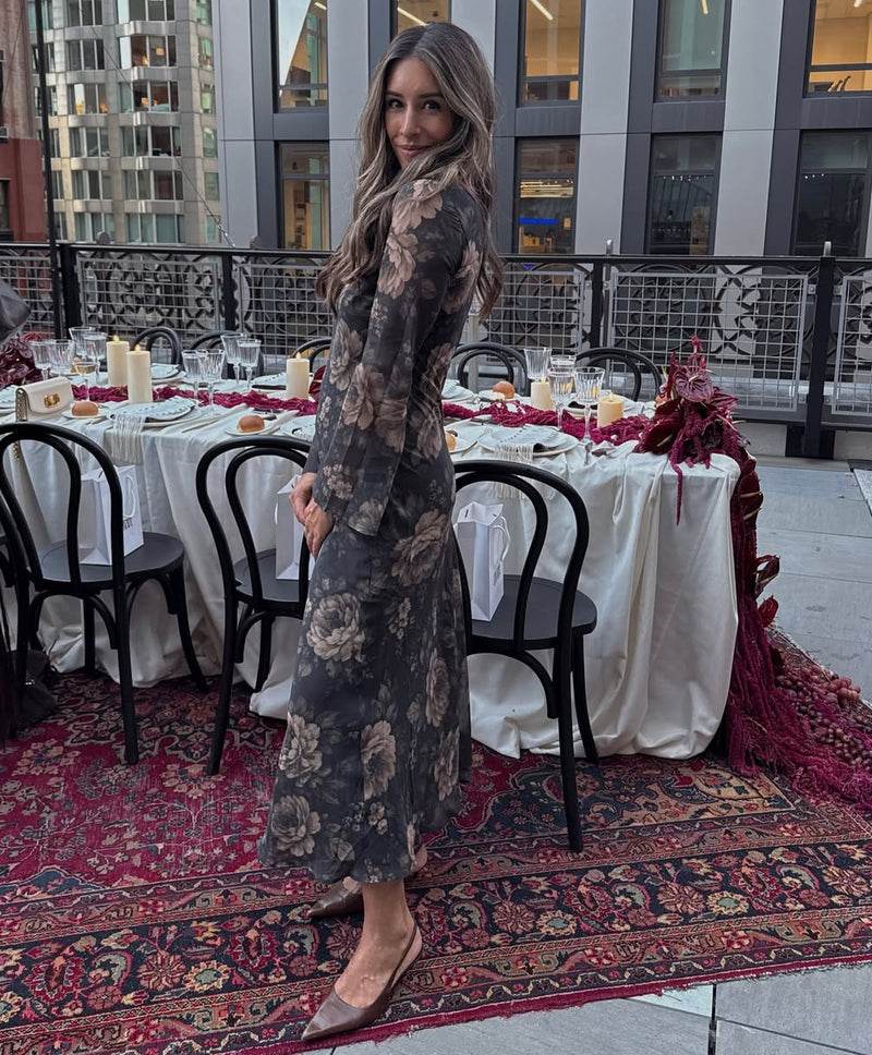 Woman wearing a long-sleeve dark gray floral maxi dress, standing on a decorated outdoor terrace set with dining tables and a patterned rug.