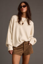 Woman wearing a cream sweater and brown shorts against a gray background
