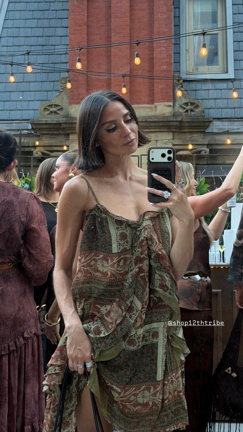 Woman taking a mirror selfie in a bohemian-style green, brown, and beige patterned dress at an outdoor evening event with string lights.